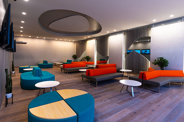 A lobby with televisions on the wall, blue and orange couches and tables.