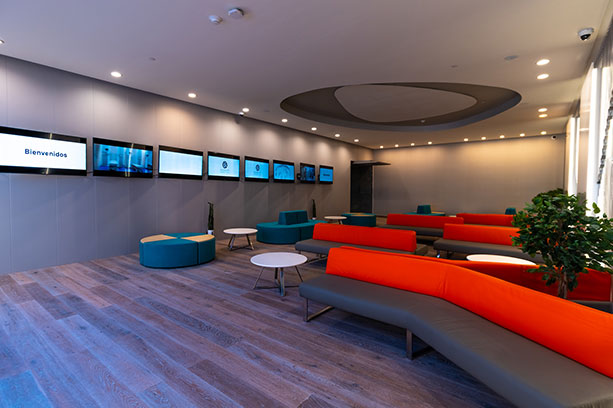 A lobby with televisions on the wall, blue and orange couches and tables.